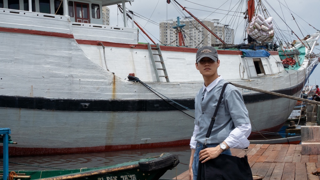 Pengunjung-mengenakan-topi-kacamata-dan-kemeja-koko-blue-grey-bertumpuk-layering-berdiri-samping-kapal-putih-tradisional-di-Sunda-Kelapa