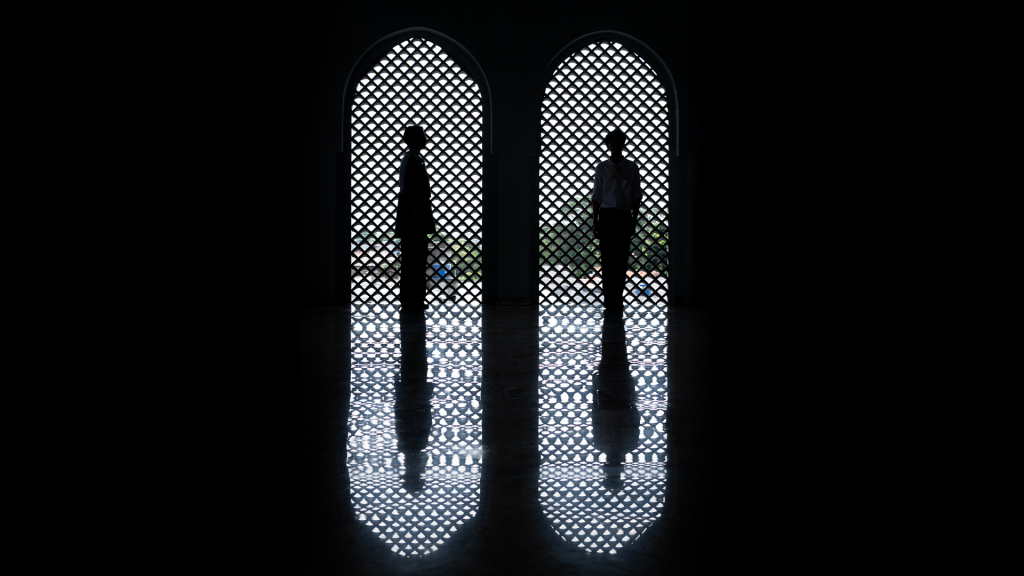 Siluet dua orang berdiri berhadapan di depan jendela melengkung dengan pola anyaman Islami, terpantul di lantai gelap yang mengilap, menciptakan komposisi simetris dan suasana tenang