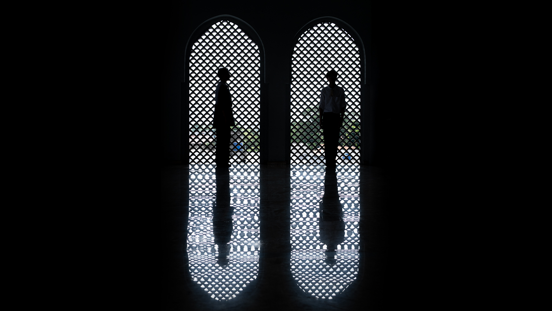 Siluet dua orang berdiri berhadapan di depan jendela melengkung dengan pola anyaman Islami, terpantul di lantai gelap yang mengilap, menciptakan komposisi simetris dan suasana tenang