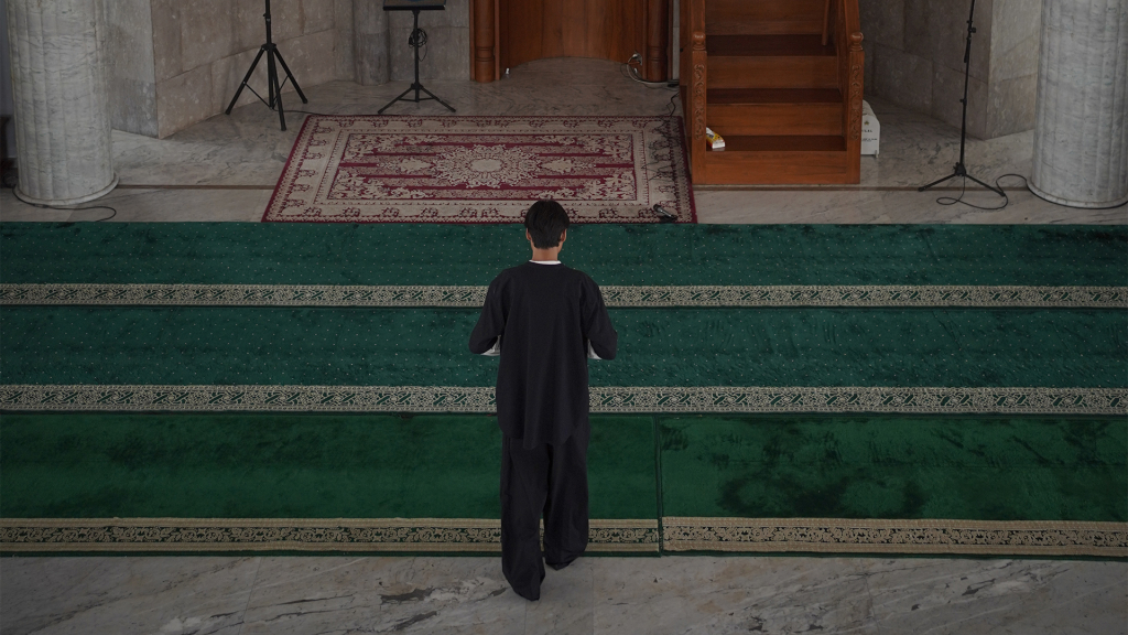 Seorang pria tampak belakang berjalan pelan di atas karpet hijau tebal masjid Pejaten Barat yang damai, menuju area mimbar kayu, lantai marmer, menciptakan suasana khusyuk dan reflektif