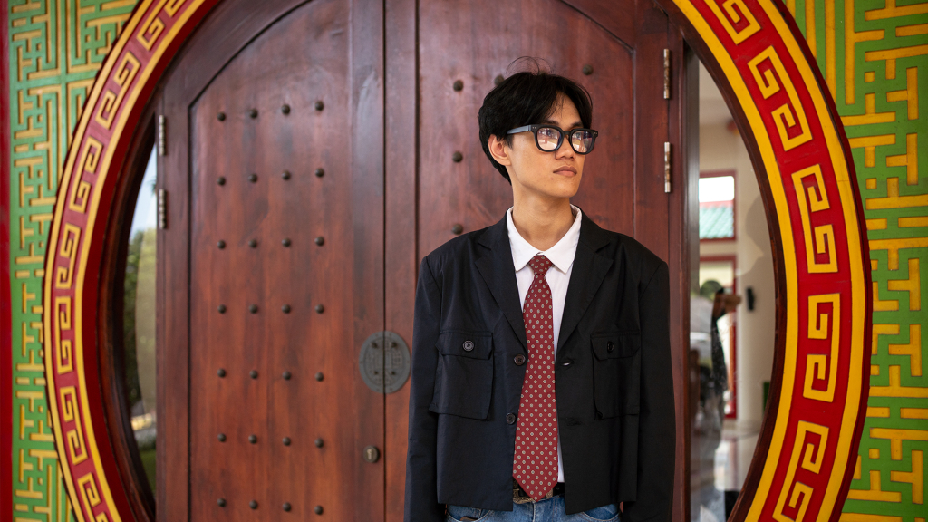 Seorang pria berkacamata dengan kemeja kerah reguler LETTI Dante putih, dasi merah, dan jas hitam, berdiri di depan pintu kayu melingkar yang dihiasi ornamen gaya Tionghoa oriental Masjid Babah Alun Desari