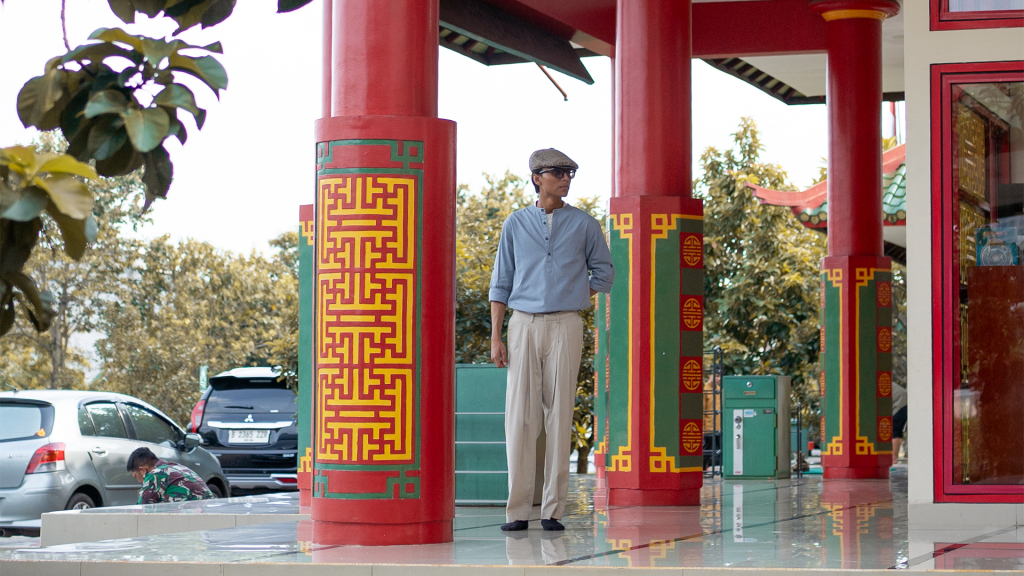 Seorang pria mengenakan kemeja biru muda, celana khaki beige, dan topi flat cap berdiri di pelataran teras dengan pilar-pilar merah besar bergaya arsitektur Tionghoa oriental Masjid Babah Alun
